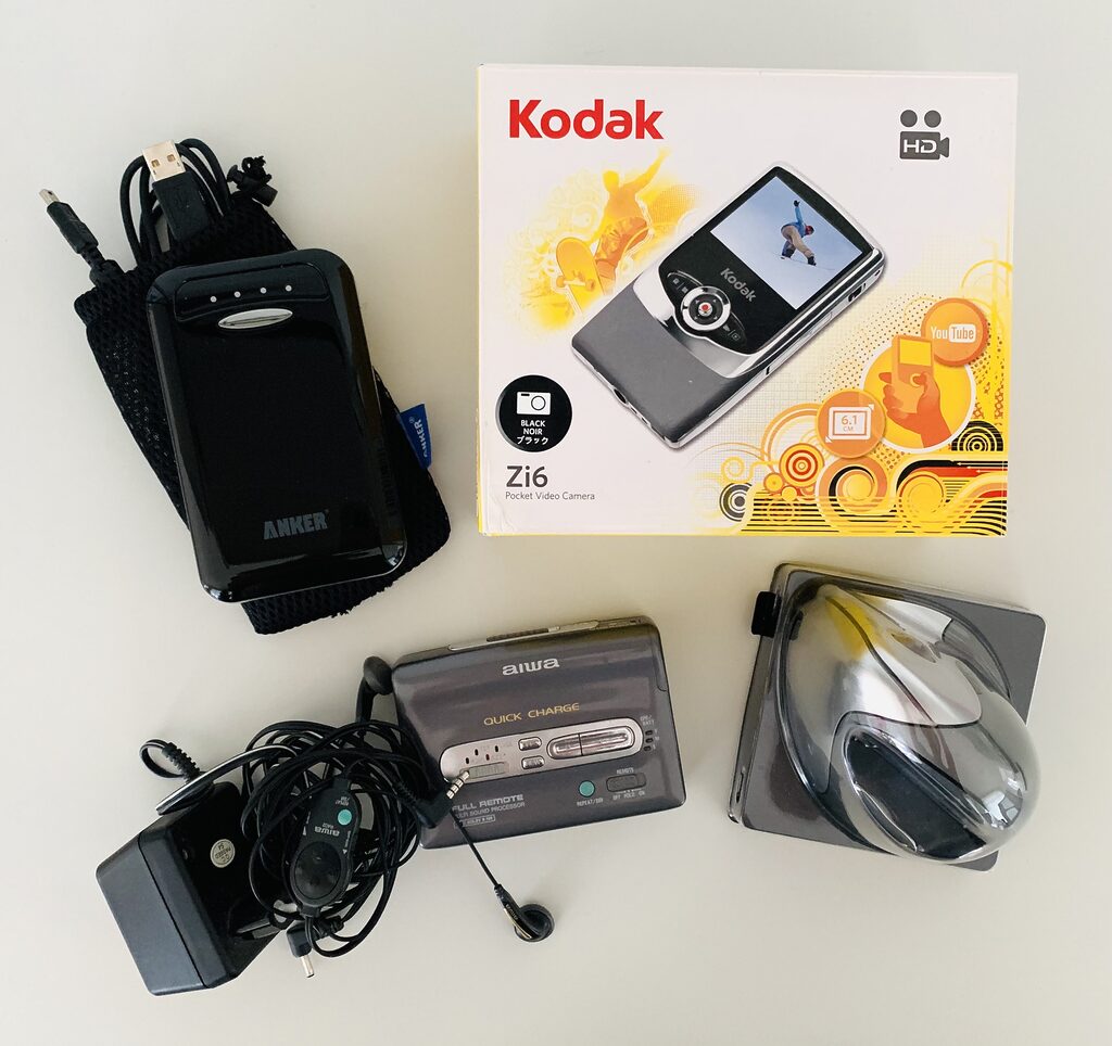Photo of an Aiwa walkman, a Kodak vlogging camera in its original box, an ergonomic mouse, and a power bank. The items are lying on a white table and photographed from above.