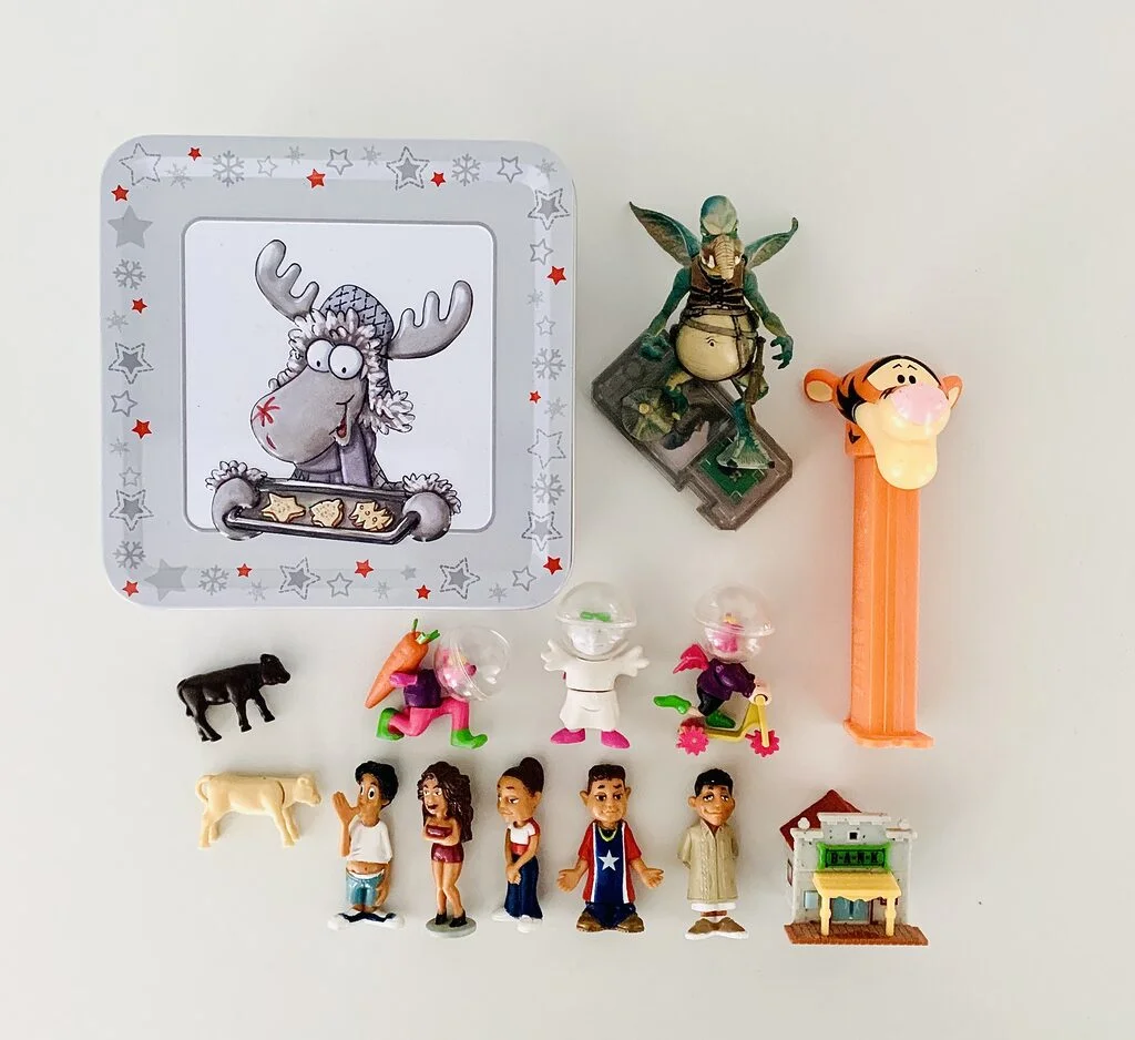 Image shows photo of decluttered, tiny toys photographed on a white table from above. Items include Kinder Egg surprises, a PEZ dispenser with Tigger the Tiger, a Star Wars figure, and little plastic animals. On the upper left is a square, light grey tin with a moose on it dressed in a Christmas outfit and holding a tray of cookies.