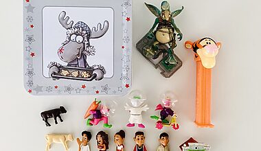 Image shows photo of decluttered, tiny toys photographed on a white table from above. Items include Kinder Egg surprises, a PEZ dispenser with Tigger the Tiger, a Star Wars figure, and little plastic animals. On the upper left is a square, light grey tin with a moose on it dressed in a Christmas outfit and holding a tray of cookies.