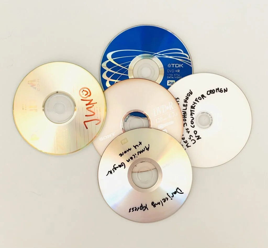 Photograph of 5 DVD-Roms without covers, which have handwritten titles on them. One of the Roms is blue. The photo is taken from above against a white background.
