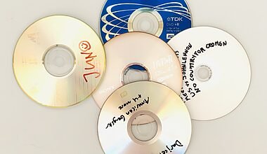 Photograph of 5 DVD-Roms without covers, which have handwritten titles on them. One of the Roms is blue. The photo is taken from above against a white background.