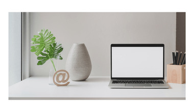 Picture of a very tidy, umcluttered white desk with a laptop open to an empty screen. To the right of the laptop is a pen holder containing pencils. To the left is an empty off-white pottery vase, next to which is a solitary green plant leaf in a slim glass vase. In front of the vases is a small wooden carving of the @ sign. The workspace appears very organized and decluttered.