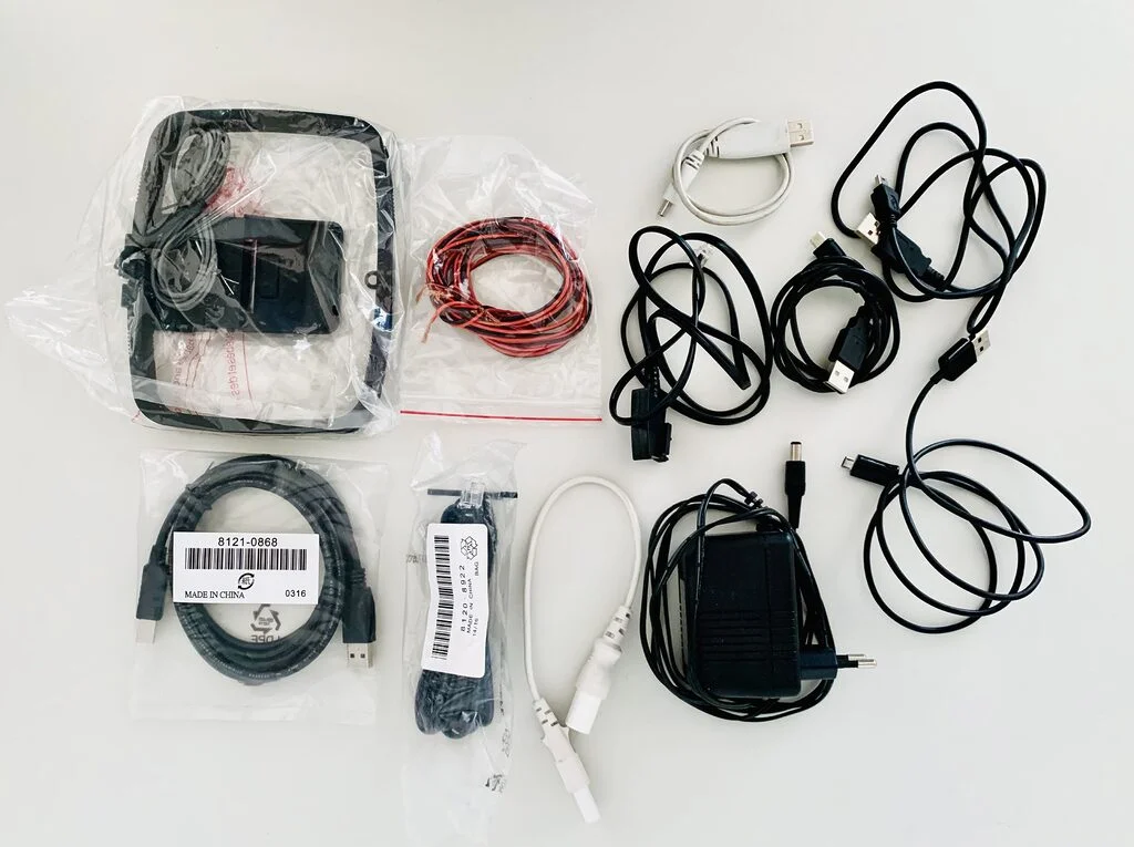 Photograph of e-waste or tech waste, laid out on a white table and photographed from above. Items include power plugs, an analog antenna, and random cables.