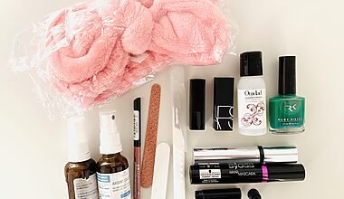 Photograph of assorted makeup and cosmetic items that were decluttered from the bathroom. Items include a big pink microfiber headband, small spray bottles, nail varnish, mascara, a nail file and lip pencil, and lipsticks. The items are neatly arranged on a white table and photographed from above.