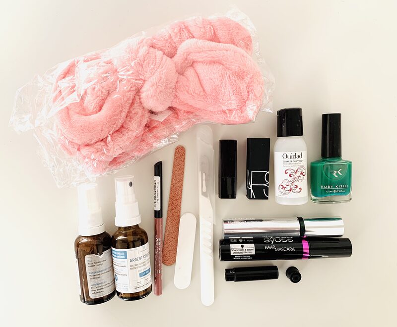 Photograph of assorted makeup and cosmetic items that were decluttered from the bathroom. Items include a big pink microfiber headband, small spray bottles, nail varnish, mascara, a nail file and lip pencil, and lipsticks. The items are neatly arranged on a white table and photographed from above.