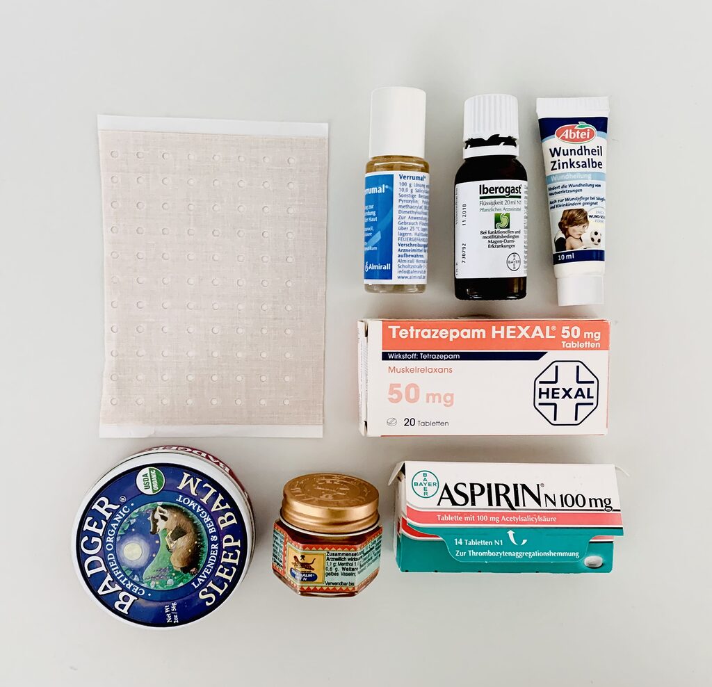 Photo of a tidy arrangement of items which were decluttered from the bathroom medicine chest. The apothecary items are arranged neatly on a white table and photographed from above. We can see a packet of expired aspirin, some zinc salve, tiger balm, a plaster and assorted drops and gels.
