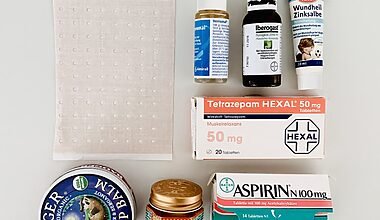 Photo of a tidy arrangement of items which were decluttered from the bathroom medicine chest. The apothecary items are arranged neatly on a white table and photographed from above. We can see a packet of expired aspirin, some zinc salve, tiger balm, a plaster and assorted drops and gels.