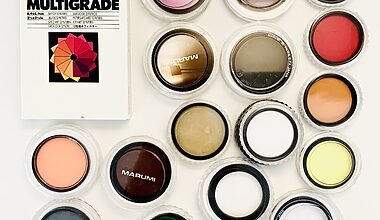 Photo of a very tidy, clean selection of new photographic filters for camera lenses in a variety of bright colours. The filters are round, 52mm to 58mm large, and in their plexiglass cases. There are about 16 such photographic filters laid out on a white table, and in the upper left is a box of Ilford Multigrade gelatine filters for the darkroom as well.