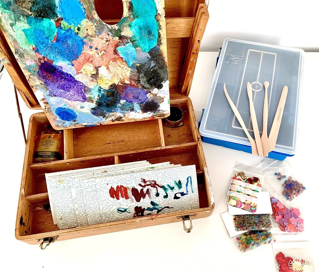 Photo of an old wooden oil paint kit from the 1960s, rather beaten up. Propped against it is the artist's palette with layers and layers of dried up, bright oil paints. Inside the kid is an empty terpentine bottle and some cut up paint by numbers boards to test the paints on. Next to the kit are some craft supplies such as beads, paper cut-outs, clay tools, and a plastic box to organise small bits in. The items are placed on a table table in front of a white wall.