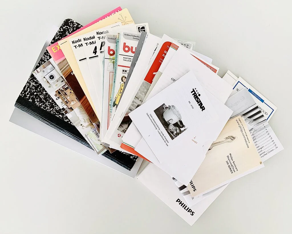 Photo of a decluttered stack of pamphlets notebooks, sewing patterns, product brochures. The stack is spread out on a white table like a fan.