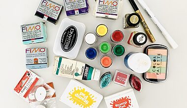 Photo of colourful craft supplies that were sorted out, including inkpads, fimo modelling clay, pens, and paints, all on a white desk.