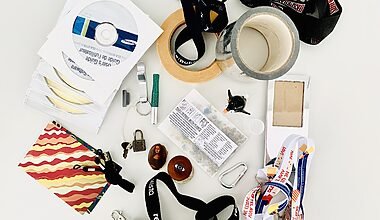Photo of around 25 pieces of random items typically found in a junk drawer such as masking tape, keychains and lanyards, old CD-Roms, a luggage lock and key, and a hook.