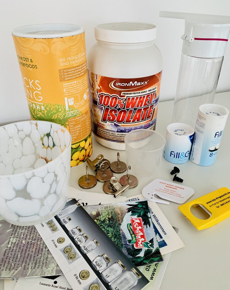 Photo of items from a kitchen clearout decluttering session, including magnets, protein powder, water filters, takeaway menus, and a bottle opener. All the objects are set on a white table in front of a white wall.