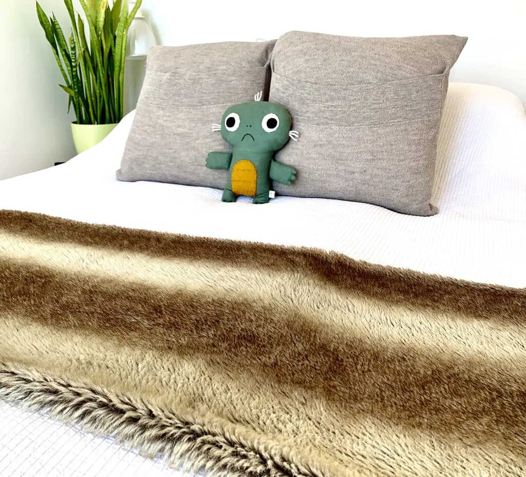 Photo of a tidily made up bed with a white bedspread, greige coloured decorative pillows, a fake fur bed throw, and a rather sad looking sort of stuffed animal. The walls are white and to the left of the bed is a tall bright green plant. Making your bed is one of the best lazy tidying tips to keep your place looking cleaner.