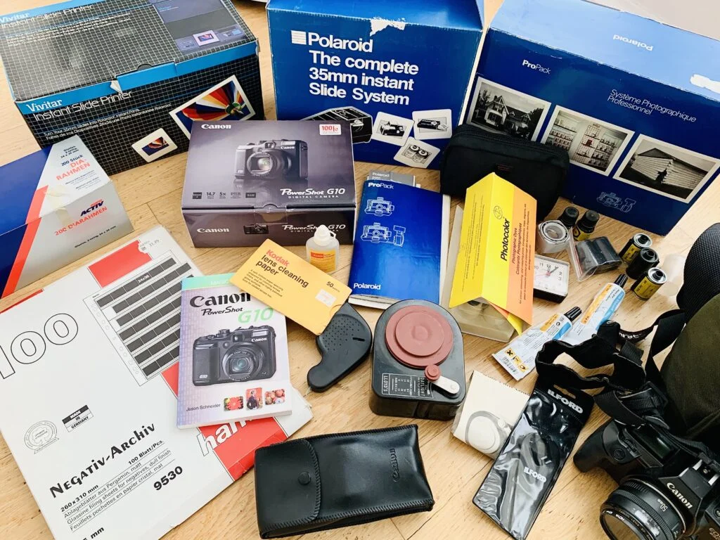 Lot of miscellaneous photographic equipment sorted out during a decluttering challenge. The items shown include analogue, Polaroid, and digital cameras, a camera bag, instruction booklets and manuals, film, and vellum envelopes for photos.