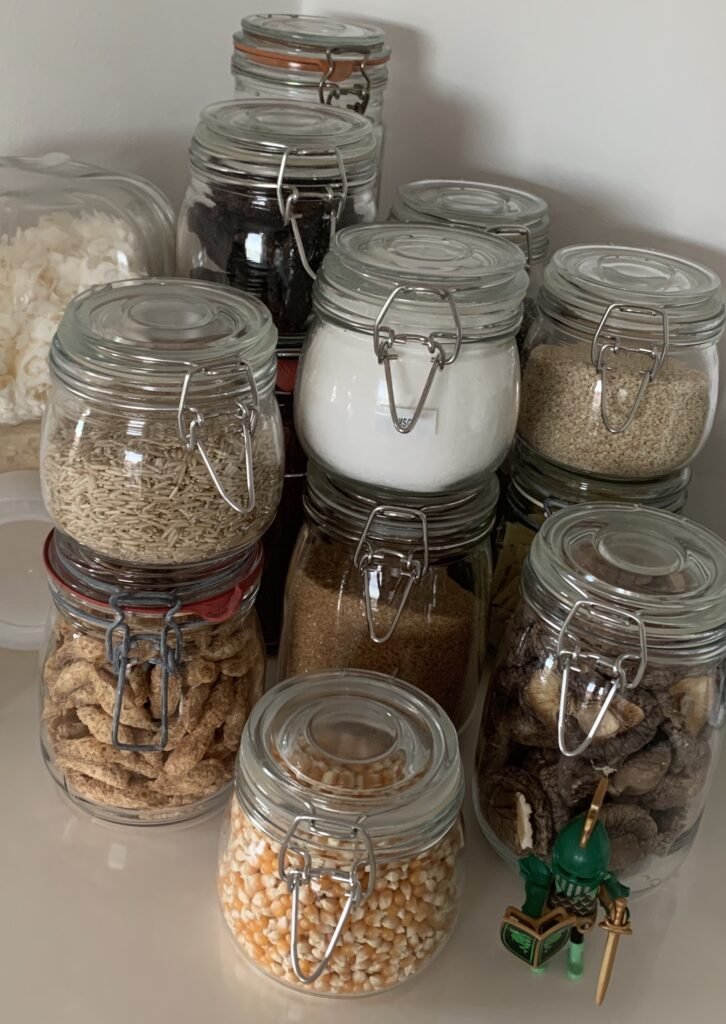 Photograph of glass jars filled with dried foodstuff like brown rice, popcorn maize, dried mushrooms, brown sugar, coconut flakes and tofu puffs. The jars are used in a small kitchen that has no pantry to maximise storage space.