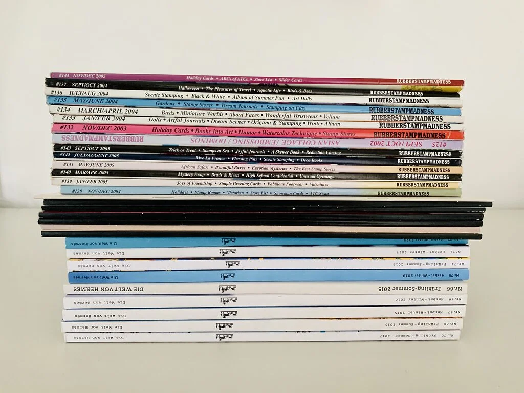 Image shows a tidy stack of decluttered magazines from the side, including issues from Hermès, Rubber Stamp Madness, and Polaroid. The stack has colourful spines and sits atop a white table in front of a white wall.