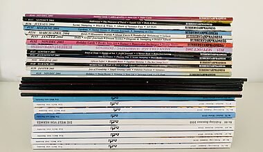 Image shows a tidy stack of decluttered magazines from the side, including issues from Hermès, Rubber Stamp Madness, and Polaroid. The stack has colourful spines and sits atop a white table in front of a white wall.