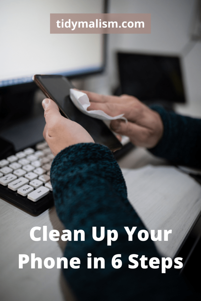 Two hands holding a smartphone at a workstation, and wiping it down with a wet disinfectant wipe. Caption reads Clean Up Your Phone in 6 Steps and is about digital decluttering of smart devices and mobile phones.