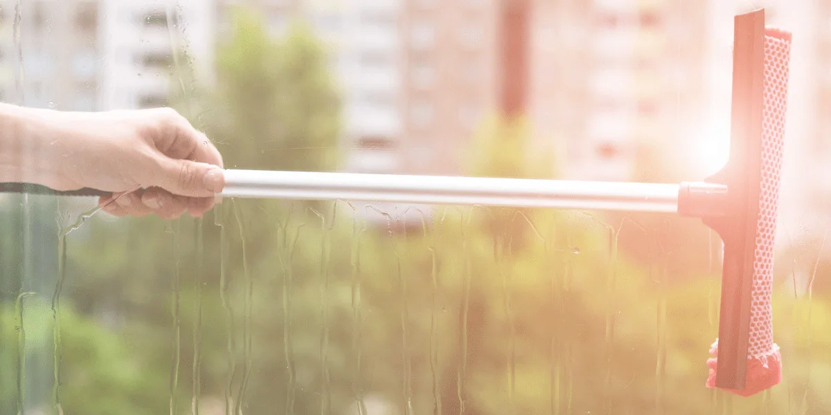 Window washing before the hot weather starts is just one thing you should have on your to-do checklist to get organised for summer. Image shows a hand pulling a squeegee across a freshly washed window. The weather looks warm and summery. The image is for a blog post about getting everything at home organised for the summer so that you can enjoy the warmer weather without worrying about chores, housekeeping, or home repairs and maintenance.