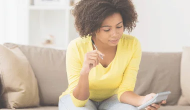 Photograph of a twenty-something black woman in a yellow long-sleeved t-shirt, sitting on her couch with a pen in one hand, and a calculator in the other.
