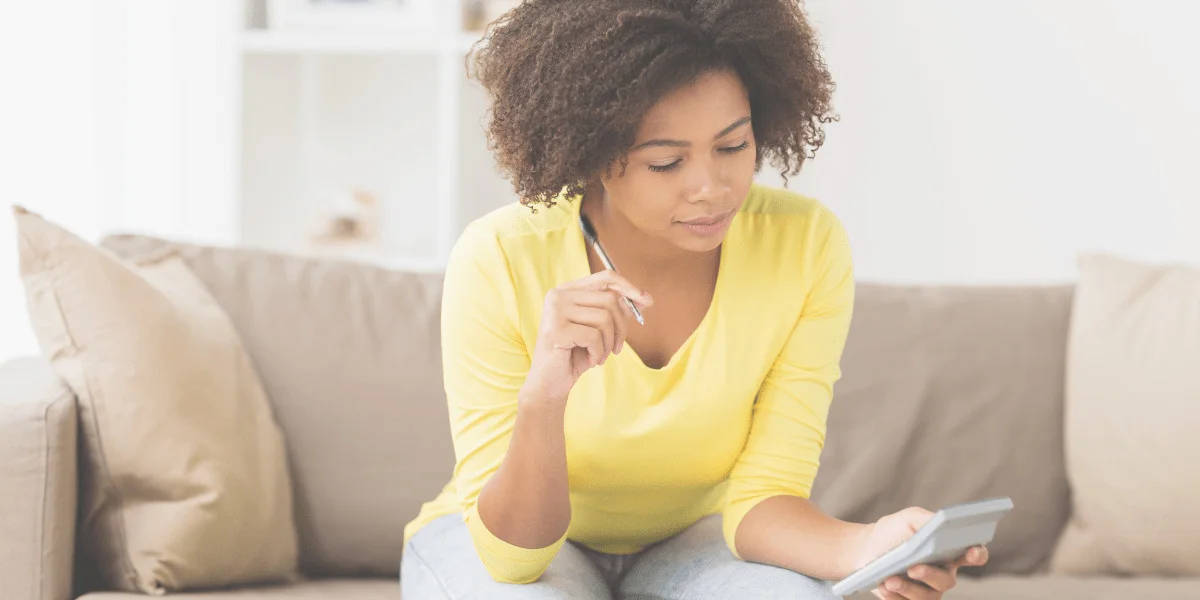 Photograph of a twenty-something black woman in a yellow long-sleeved t-shirt, sitting on her couch with a pen in one hand, and a calculator in the other.