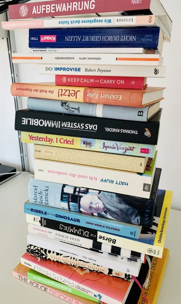 A stack of German and English books piled high on a white desk.