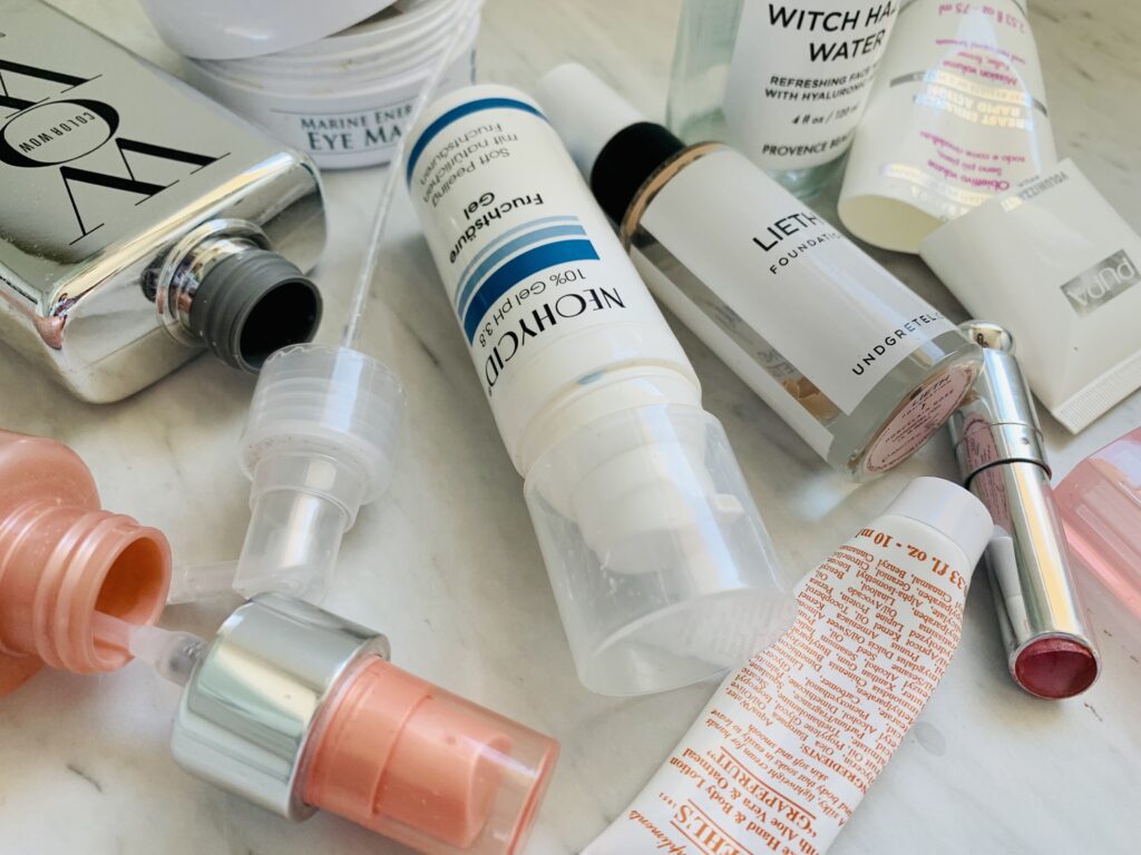 Close up photograph of an assortment of empty beauty products used up during a low buy challenge. Tubes, plastic vials, glass bottles all randomly lying about on a marble countertop. Some of the bottles are open and lying on their side. Some of the tubes are cut in half and we can see that the remainig product was really used up.