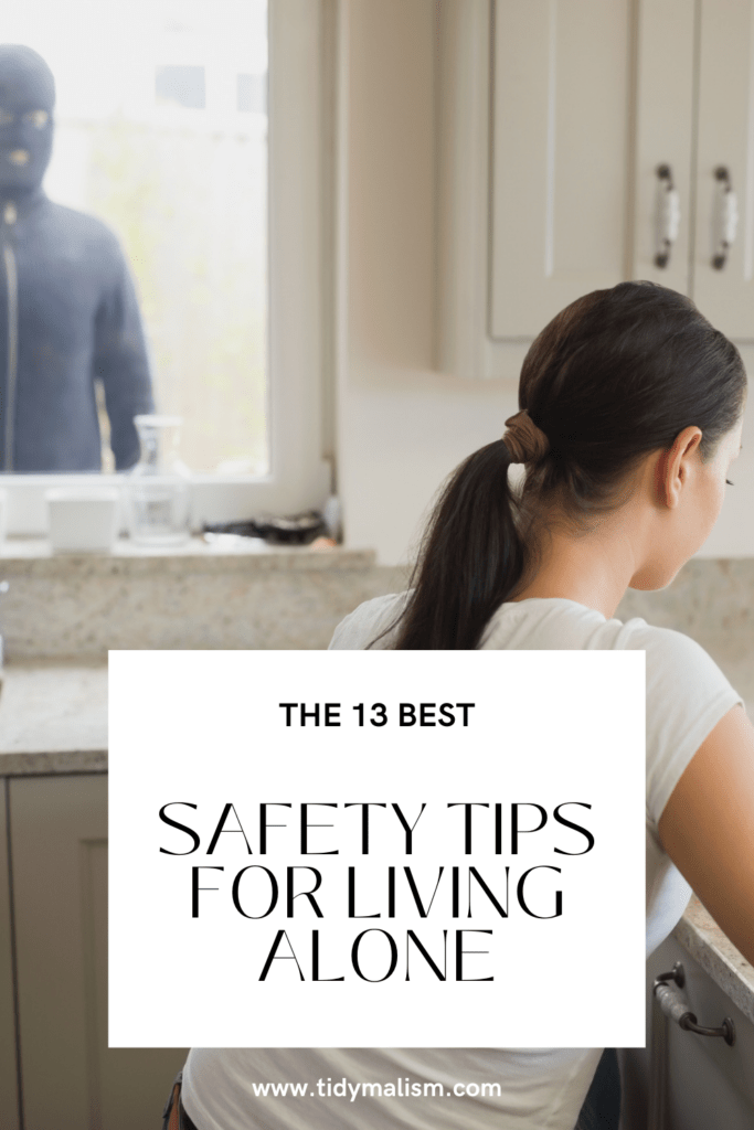 Young lady sitting at her countertop on her laptop. At the window beyond her left shoulder, we can see an intruder spying on her. He is wearing black and a black ski mask. Caption reads The 13 Best Safety Tips for Living Alone.