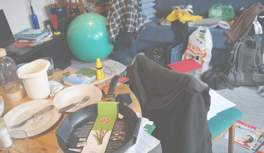 Messy, cluttered and dirty room, with items strewn everywhere and dirty dishes on the dining table.