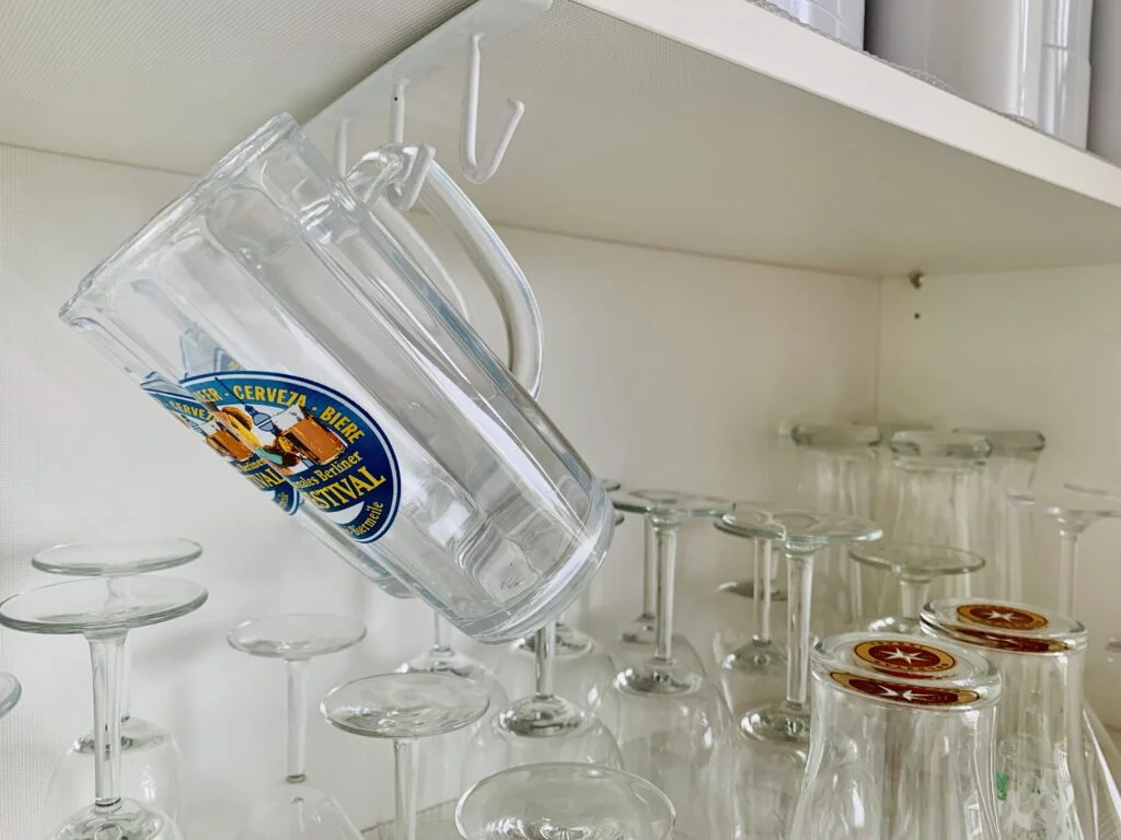 Inside of an upper kitchen cabinet used to store glasses. We can see wine glasses, beer glasses, champagne glasses, and martini glasses. There is a row of hooks inside the upper shelf with glass beer mugs hanging to maximize space in a small kitchen.
