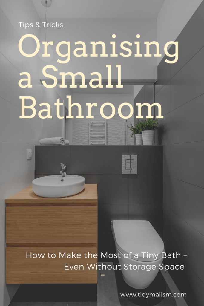 Photograph of a very tiny, yet very tidy bathroom. The walls and floor are done in dark grey, large-format tiles. The toilet is a hanging version in white ceramic with plumbing built into the wall. The white ceramic sink sits atop a modern, square oakwood cabinet. Caption reads tips & tricks for organising a small bathroom. How to make the most of a tiny bath, even without storage space. tidymalism.com