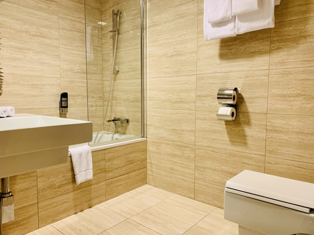 Photograph of a hotel bathroom. There is a bathtub with glass shower divider, chrome fixtures, large sink, and modern toilet bowl. The walls and floor are done in large-format beige colored stone tiles, and the porcelain fixtures are all white, as are the fluffy bathtowels.