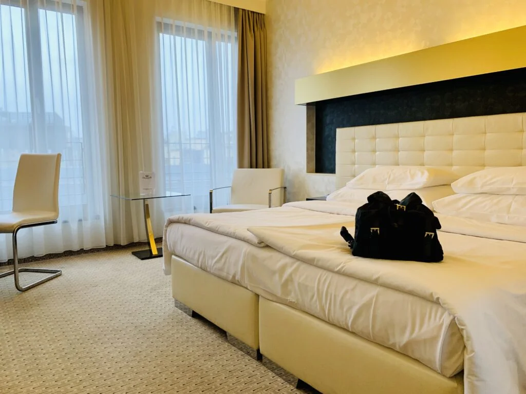 Photo of very organised hotel room. The bed is neatly made and there is a small black Prada backpack on top of the bed.