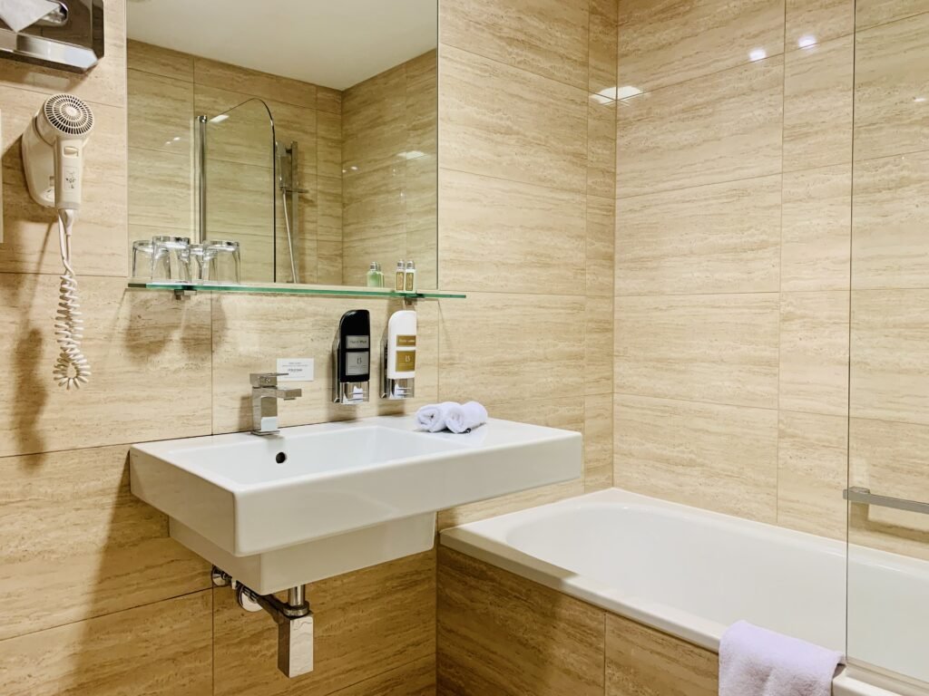 Photograph of an immaculately clean bathroom in a hotel room, with a large sized sink, mirror, and bathtub. There is a hairdryer installed on the wall to the left of the sink, as well as a chrome box of tissues.