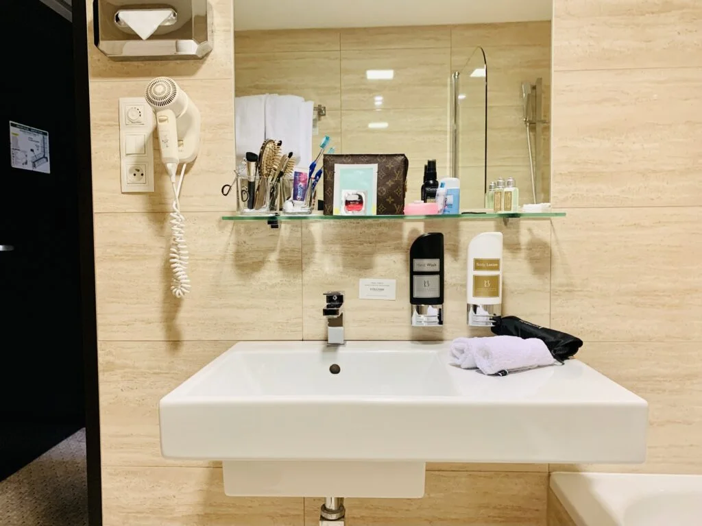 Photograph of a hotel bathroom sink after the guest has unpacked her toiletries. The space above and around the sink is very organized and tidy.