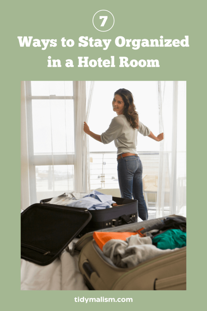 Photograph of a young lady who just checked into her hotel room. Her two carry on suitcases are lying on the bed opened, but not yet unpacked. She's opening the curtains beyond the bed and turned around to smile at the camera. Caption reads: 7 ways to stay organized in a hotel room.