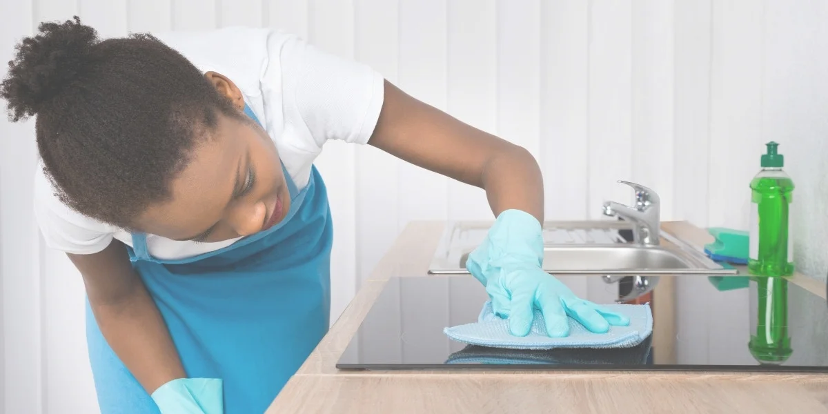 Young black woman in a white t-shirt and bright blue apron with her hair tied back, and wearing light blue rubber gloves. She's bending over her ceran stovetop and wiping it down.