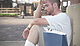 Photograph of a twenty-something white male sitting on the street corner in shorts and t-shirt. He looks really disappointed and holds his palm to his forehead. To his left is a large shopping bag.