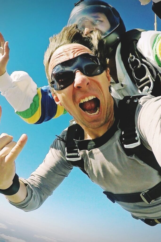 Photograph of a thirty-something guy doing a tandem skydiving jump. He's strapped onto his tandem partner high in the sky, and screaming of joy as he takes a selfie. Skydiving vouchers are great minimalist gift ideas.