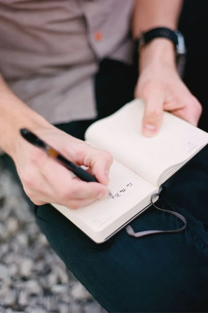 Cropped photo of two white hands making a note of something in a small-sized Moleskin cahier notebook.