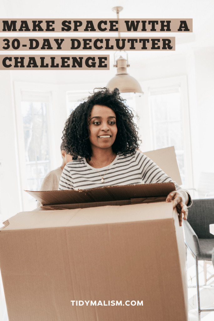 Pretty lady with dark curly hair, wearing a white long-sleeved top with navy stripes. She's carrying a box out of her sunny and bright living room, it looks as if she's decluttering stuff. Caption reads make space with a 30-day declutter challenge.