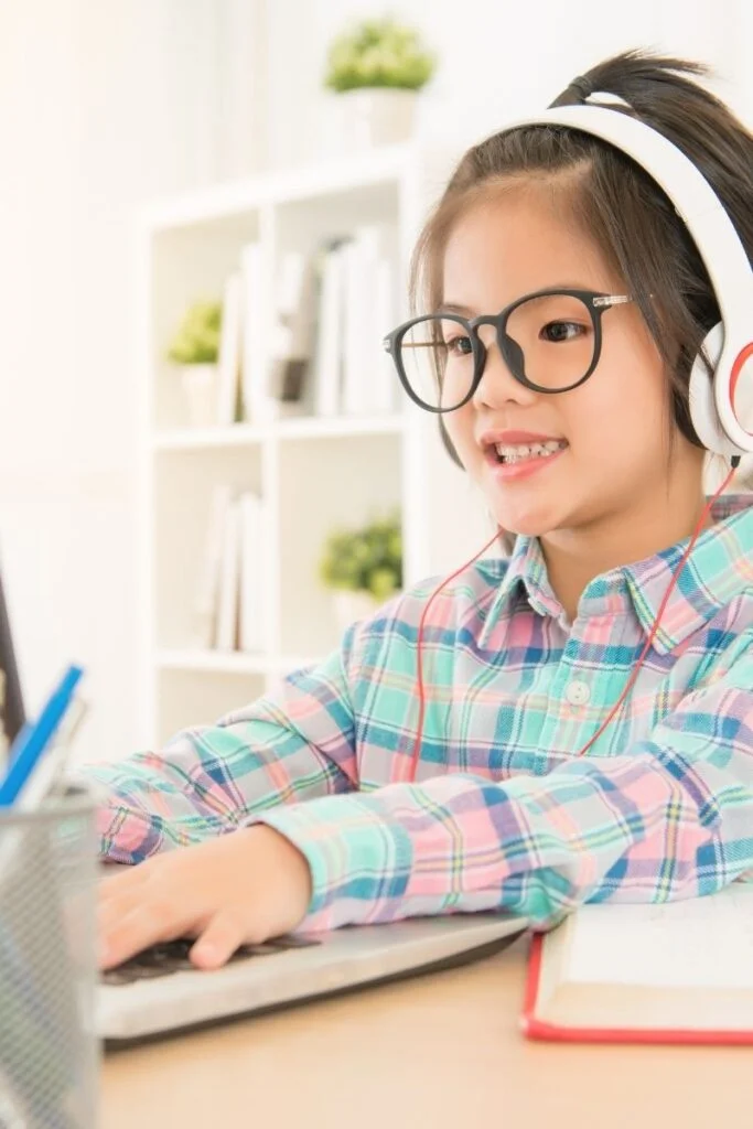 Photograph of a little girl with dark hair and glasses, sitting at a laptop with both arms stretched over the keyboard. She's wearing white headphones and a pastel checked shirt and looks very engaged in what she's learning. She's smiling straight on at the laptop.