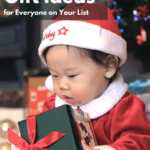 Asian baby dressed in a red and white Santa outfit, sitting upright holding a small gift box in his or her hands. Behind the baby, we can make out a blurry Christmas tree and tinsel in the background. Caption reads Minimalist Gift Ideas for Everyone on Your List.