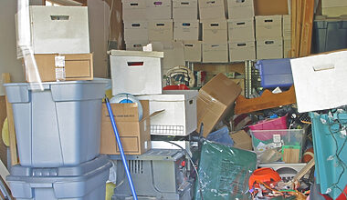 Photograph of a very cluttered garage with numerous brown moving boxes stacked up against the back wall, and loads of bags, other boxes, and household junk cluttering the entire space.