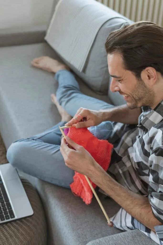 Photograph of a white male in his thirties sitting barefoot on his couch in trousers and a flanner shirt. His laptop is open to his left on the coffee table but he is busy knitting a sweater with red yarn and large knitting needles. He's smiling whilst knitting, apparently enjoying his hobby.