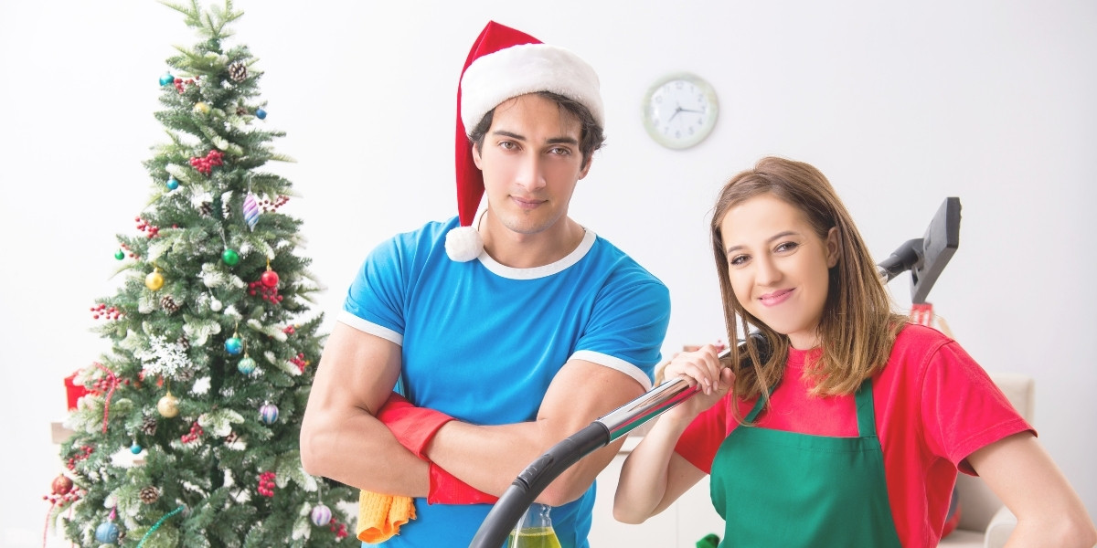 Photograph of a twenty-something couple dressed in bright, Christmas-colored clothing. The guy is wearing a Santa hat and they're tryig to get things cleaned up in front of the Christmas tree.