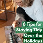 Middle-aged white woman with a blond-grey bob and glasses leaning over a box of Christmas tree lights on the floor, either removing the lights or putting them back in. Caption reads: 6 tips for staying tidy over the holidays.