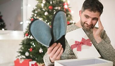 Photo of a thirty-something white male sitting in front of a Christmas tree with an opened gift box on his lap. He has pulled out a pair of drab grey slippers with his right hand, and touches his temple with his left hand with a facial expresssion of slight dismay as he holds up the gift.