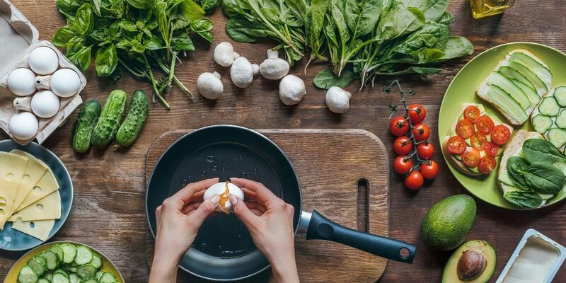 Photo taken from above of two hands cracking an egg over a skillet on a wooden countertop. Around the skillet we can see healthy green leafy vegetables, garlic, and mushrooms.